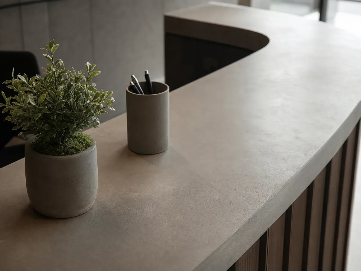 A curved concrete-effect reception desk holds a small potted plant and a cylindrical pen holder containing several pencils, with wooden panelling visible below the counter edge