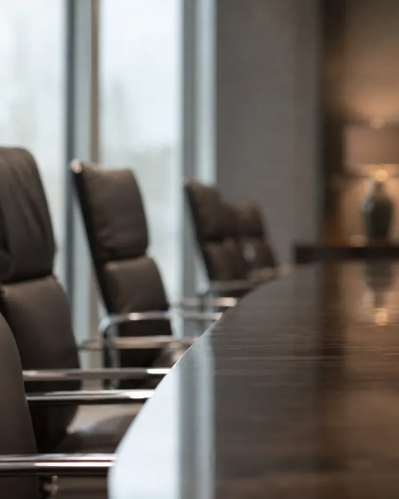 An empty boardroom with a row of dark leather executive chairs along a polished conference table, large windows behind and a softly blurred seating area beyond